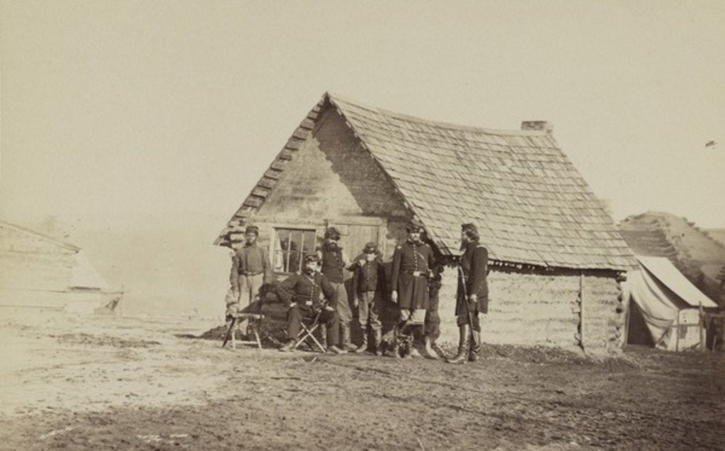 Detail of A group of soldiers, and two young men, one an African American, stand outside of log cabin quarters by American Photographer
