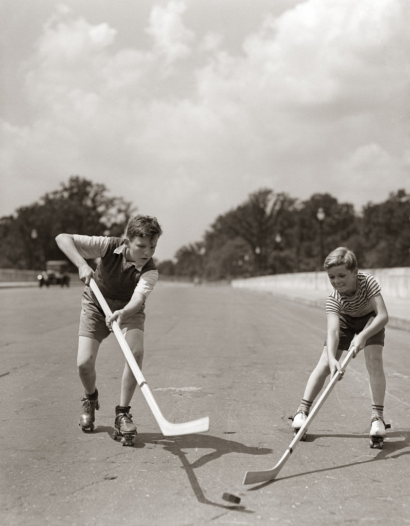 Detail of 1930s 1940s 2 Boys With Sticks And Puck Wearing Roller Skates Playing Street Hockey by Corbis