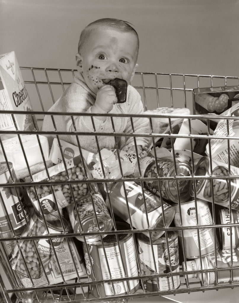 Detail of 1960s Baby Sitting In Supermarket Cart Full Of Cans Eating Candy Bar With A Messy Face by Anonymous