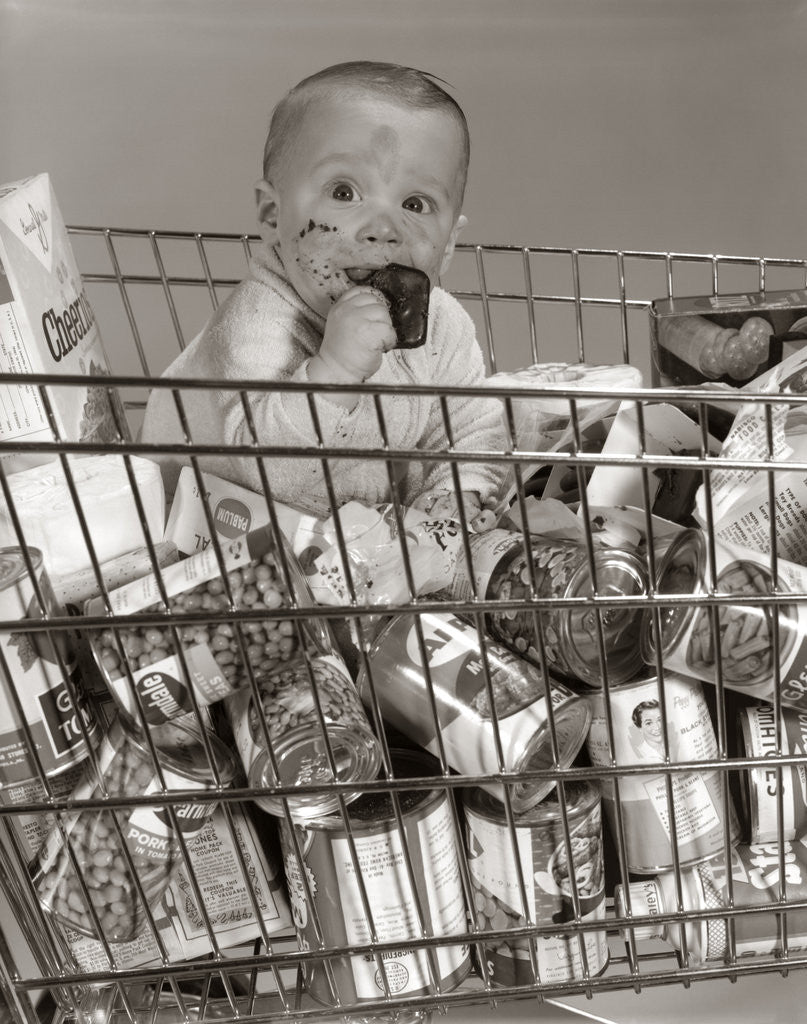 Detail of 1960s Baby Sitting In Supermarket Cart Full Of Cans Eating Candy Bar With A Messy Face by Corbis