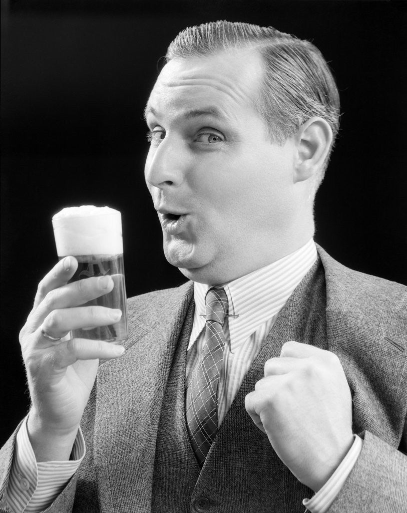 Detail of 1930s Man Holding Full Foamy Glass Of Beer Making A Funny Face With His Thumb Hooked Confidently Under His Vest by Corbis