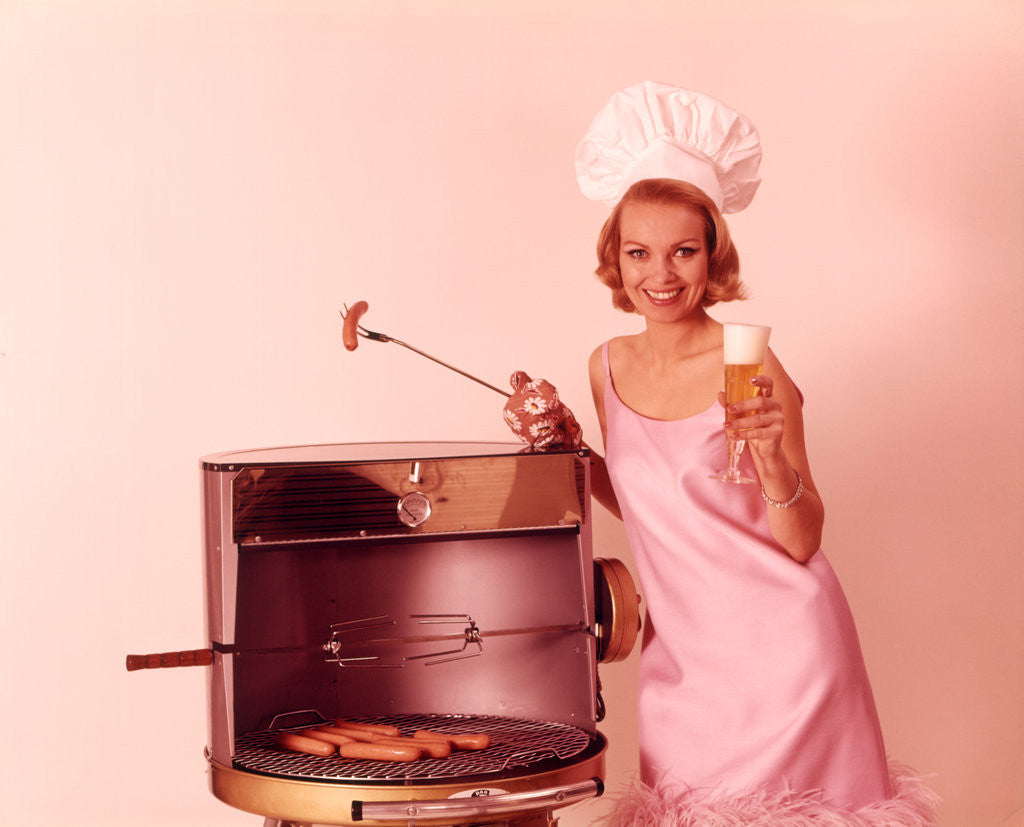 Detail of 1960s Woman Wearing Pink Party Dress And Chef Hat Grilling Hot Dogs Drinking Beer by Corbis