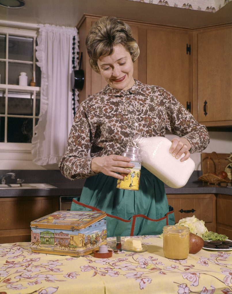 Detail of 1960s Woman Housewife Mother Wearing Apron In Kitchen Pouring Milk Into Thermos For School Lunch by Anonymous