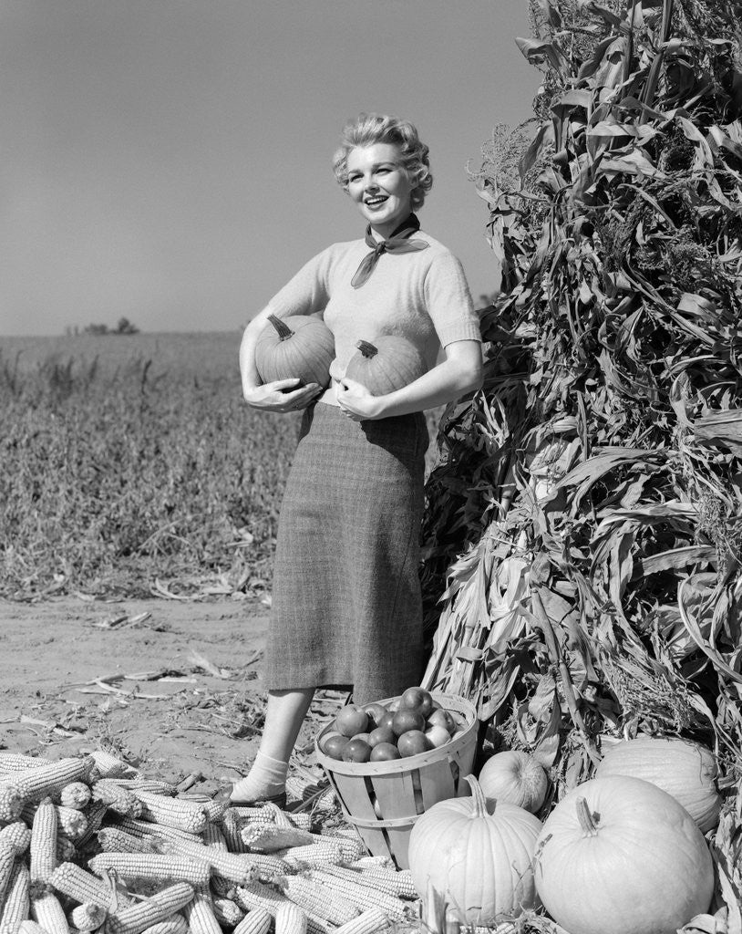 Detail of 1950s 1960s Woman Standing By Corn Shock Holding A Pumpkin Under Each Arm by Corbis