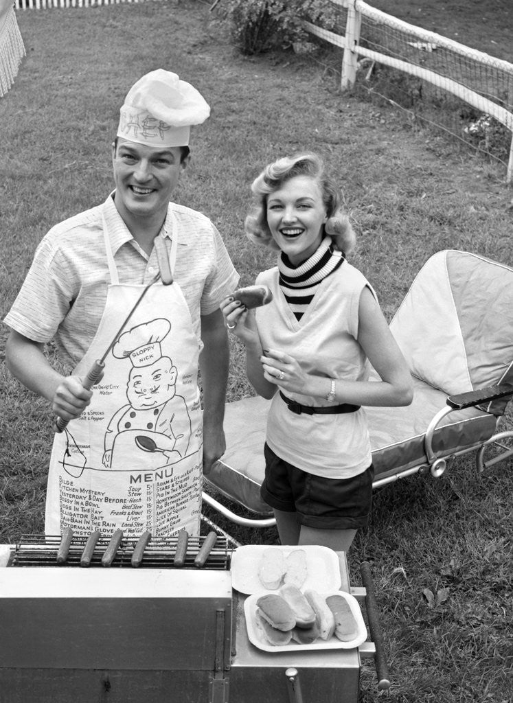 Detail of 1950s Couple Backyard Grill Cook Hot Dogs Man Wearing Apron Toque and Skewered Hot Dog by Corbis