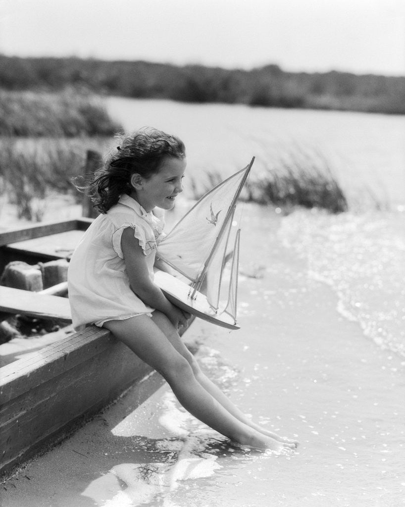 Detail of 1930s Young Girl At Seashore Holding Sailboat Toy Sitting On Edge Of Rowboat Feet In Water by Corbis