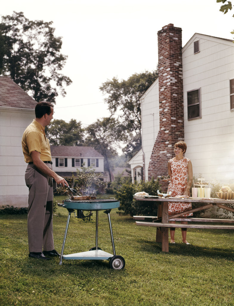Detail of 1960s Couple Suburban Backyard Man Grilling Woman Setting Picnic Table by Anonymous