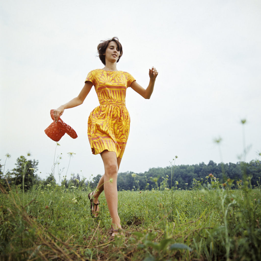 Detail of 1970s Young Woman Wearing Colorful Print Dress Running In Field Carrying Red Straw Hat by Anonymous