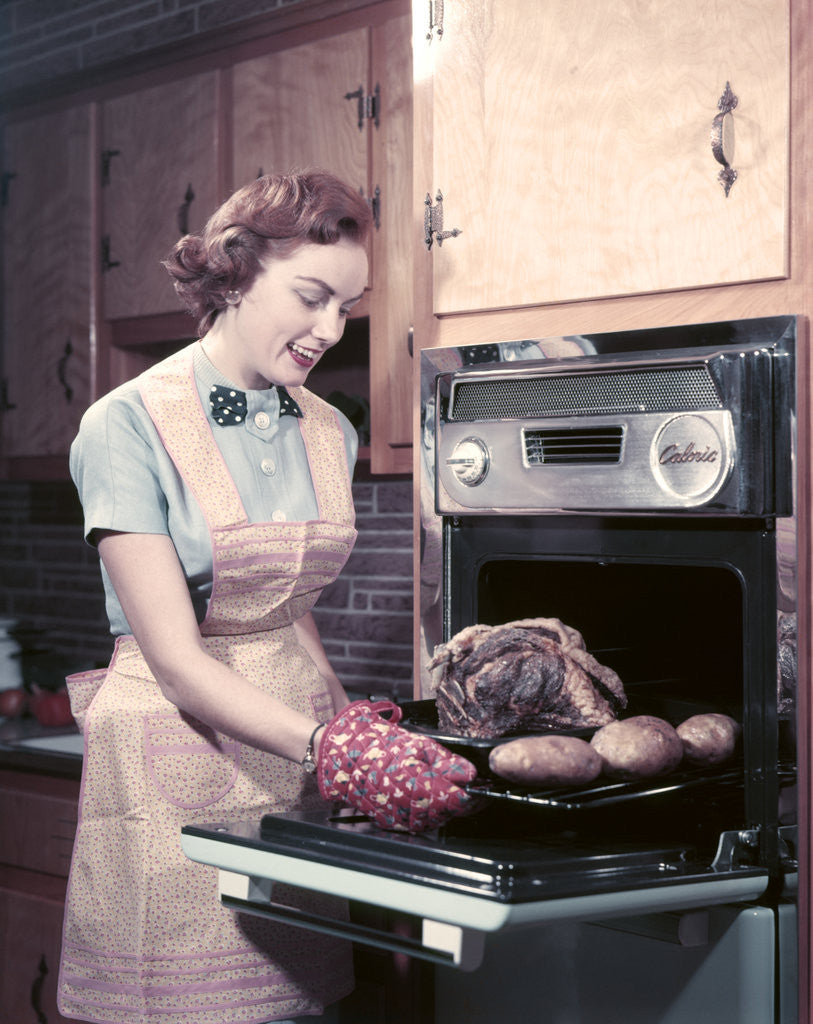 Detail of 1950s Housewife Wearing Apron And Oven Mitts Taking Roast Beef With Potatoes Out Of Kitchen Oven by Corbis