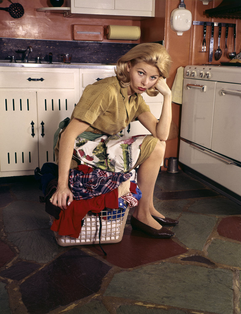 Detail of 1960s Weary Dejected Woman Housewife Homemaker Sitting On Full Laundry Basket In Kitchen by Anonymous