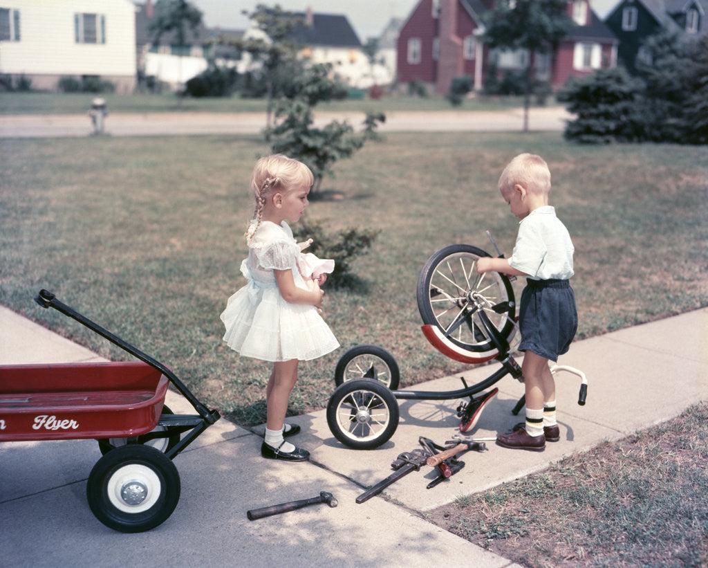 Detail of 1950s Little Girl Sister Holding Doll Watching Little Boy Brother Repair Tricycle by Corbis