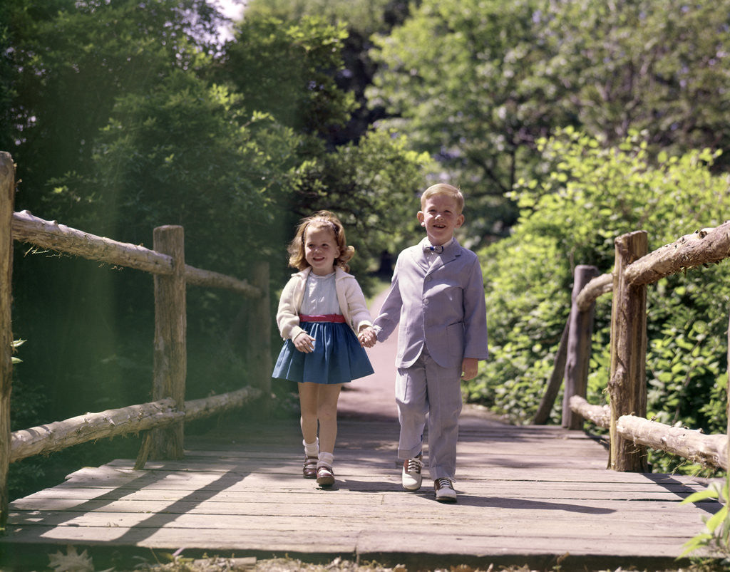 Detail of 1960s Little Boy And Girl Holding Hands And Walking Across Wooden Footbridge by Corbis