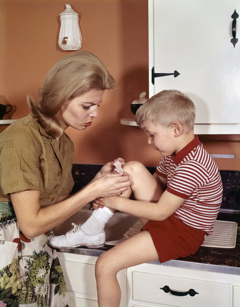 Detail of 1970s Mother Giving First-Aid Putting Band-Aid On Knee Of Son In Kitchen by Anonymous