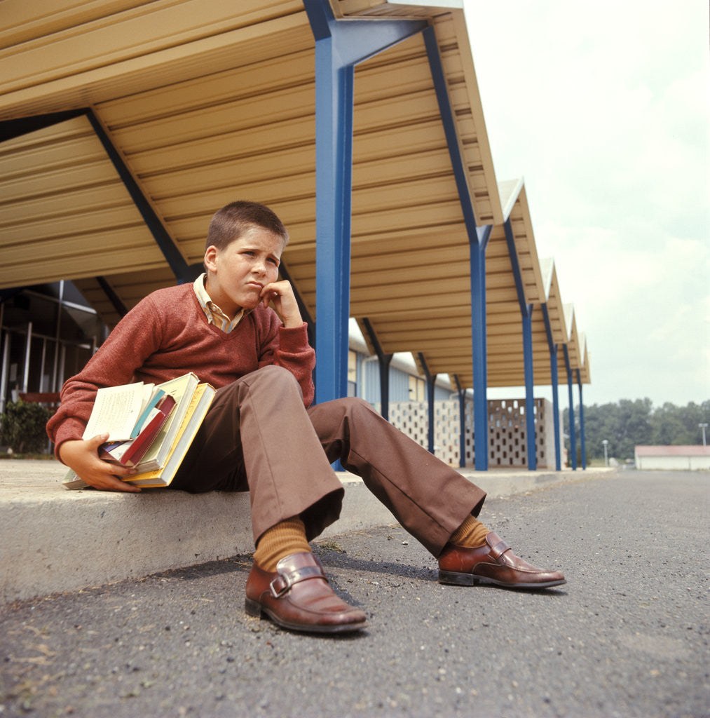 Detail of 1970s Unhappy Worried School Boy Holding Books Sitting On Sidewalk Curb Outside School Building by Anonymous