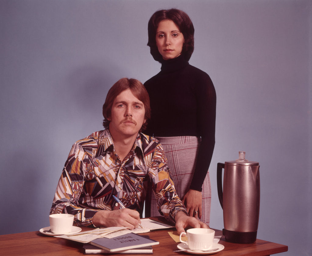 Detail of 1970s Husband Wearing Loud Print Shirt Writing Family Budget and Wife Standing Behind Coffee Pot by Corbis