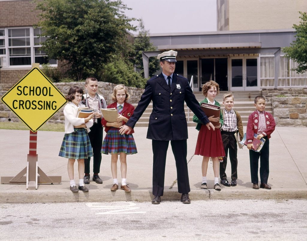 1960s Police Officer Holding Back Elementary School Children Waiting At Curb To Cross Street by Corbis