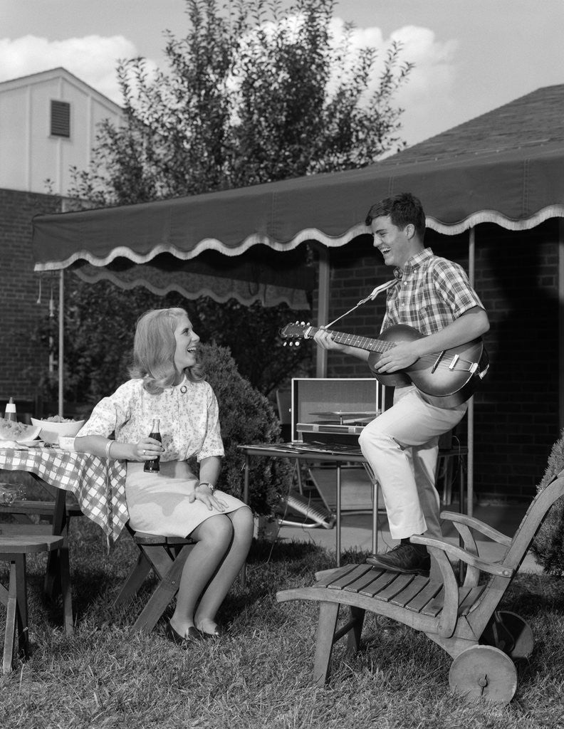 Detail of 1960s Boy Playing Guitar, Woman Holding Soft Drink Bottle Outside In Backyard by Anonymous