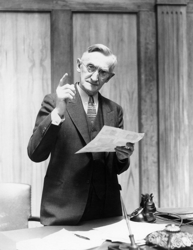 Detail of 1930s Elderly Man In Office Standing Behind Desk Gesturing With Raised Finger by Anonymous