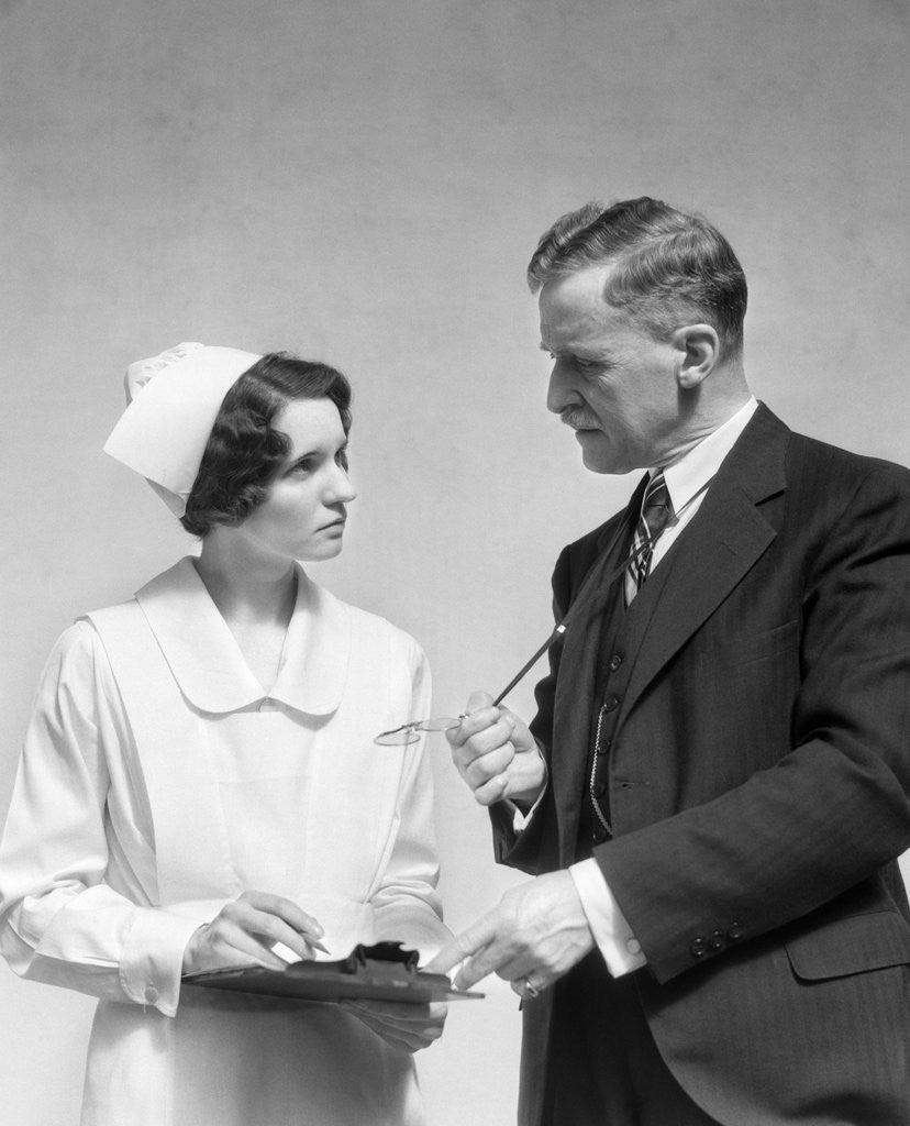 Detail of 1930s Doctor In Suit And Necktie Confers With Nurse In Uniform And Cap Holding Clipboard Chart by Corbis