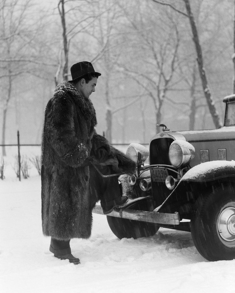 Detail of 1930s Man In Hat And Raccoon Fur Coat Standing Foot On Bumper Of Chevrolet Roadster Stalled In Snow Storm by Anonymous