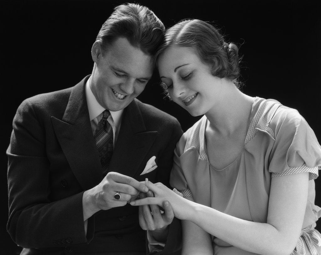 Detail of 1930s Couple Sitting Heads Pressed Together Man Putting Engagement Ring On Woman's Finger by Corbis