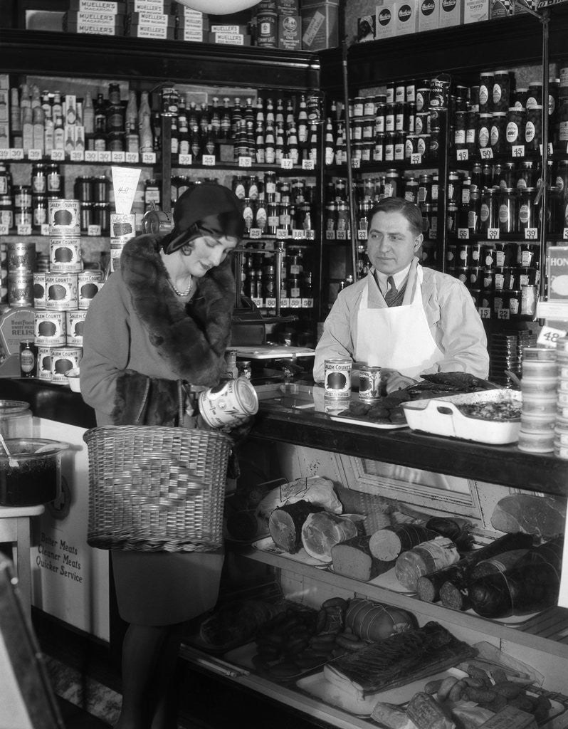 Detail of 1920s Store Clerk Watching Woman Grocery Shopping Reading Label On Canned Goods by Corbis