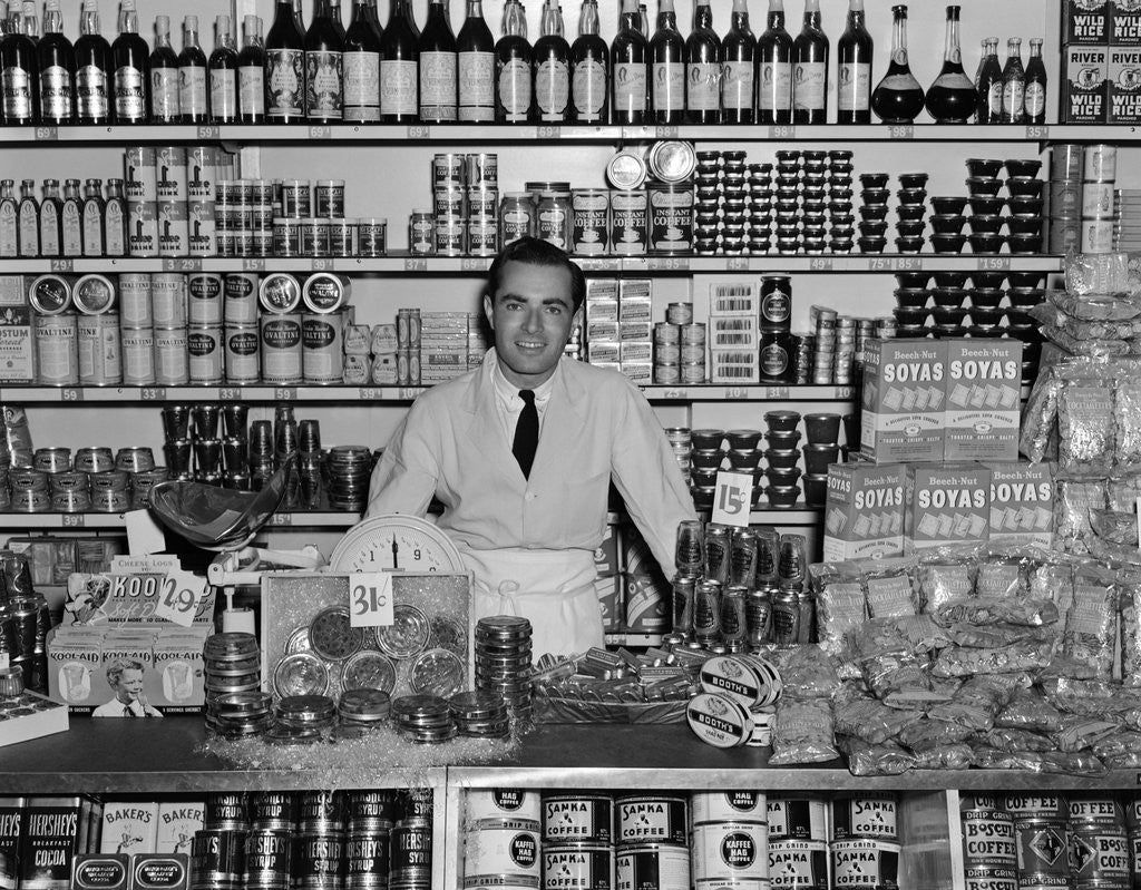 Detail of 1940s Grocer Standing Behind Counter Filled With Various Food Products by Anonymous