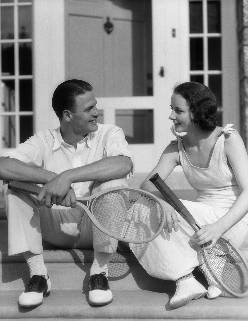 Detail of 1930s Couple Man Woman In Tennis Whites Holding Racquets Sitting Talking On Steps In Front Of House Outdoor by Anonymous