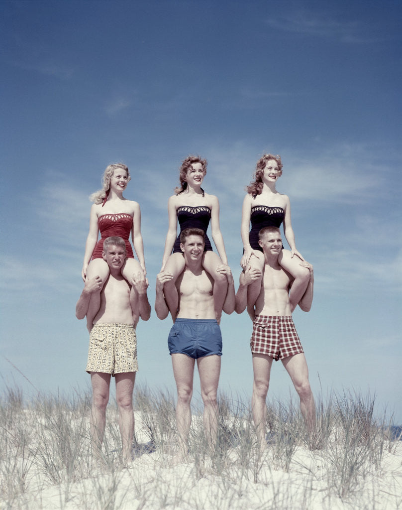 Detail of 1950s 3 Teen Couples Standing On Sand Dune Girls Sitting On Guys Shoulders by Corbis