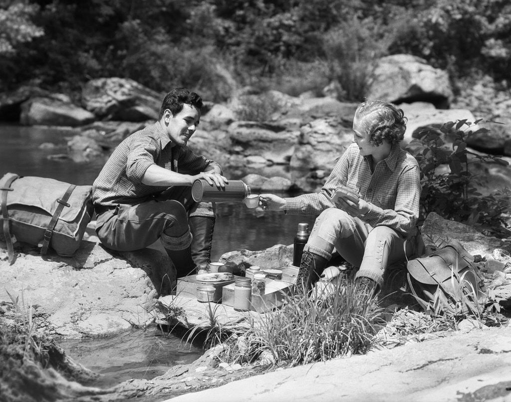 Detail of Couple having picnic sitting on rocks by stream man pouring drink for woman from thermos by Anonymous