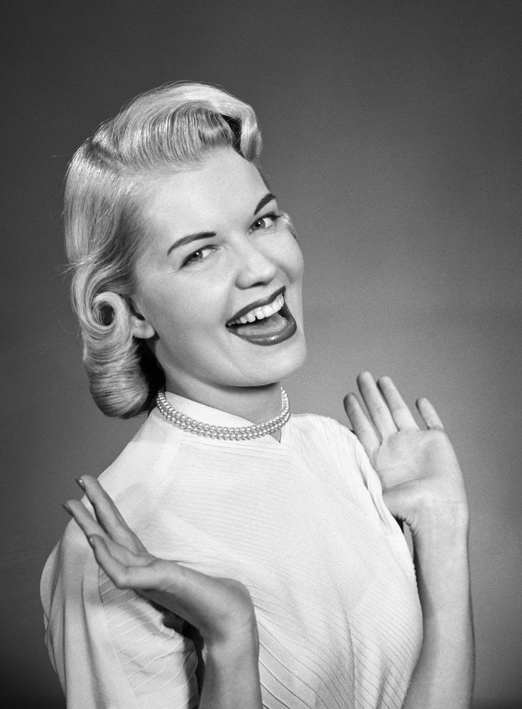Detail of Woman wearing pearls smiling with hands up thumbs tucked under arm by Anonymous