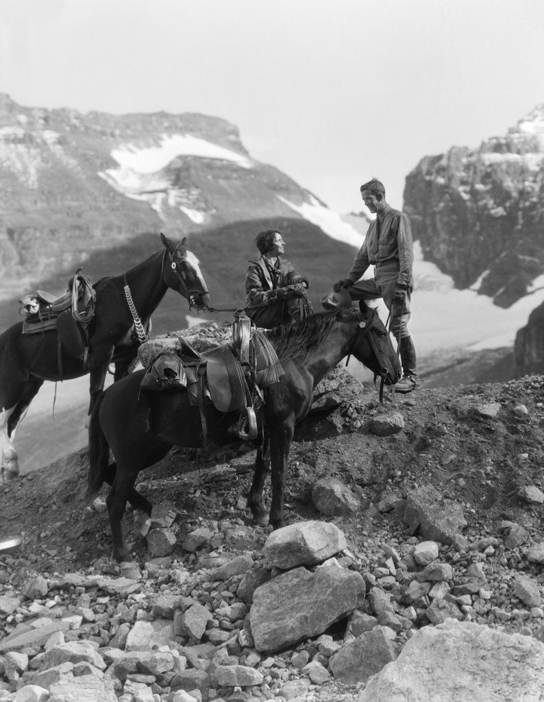 Detail of Couple man woman wearing riding gear jodhpurs boots spurs sitting standing on large rock by two horses by Anonymous