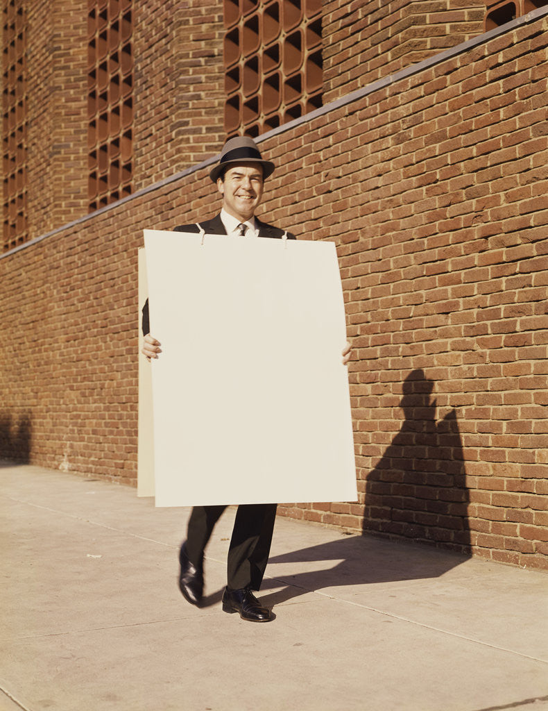 Detail of Smiling man wearing suit necktie and hat walking carrying large sandwich board by Anonymous