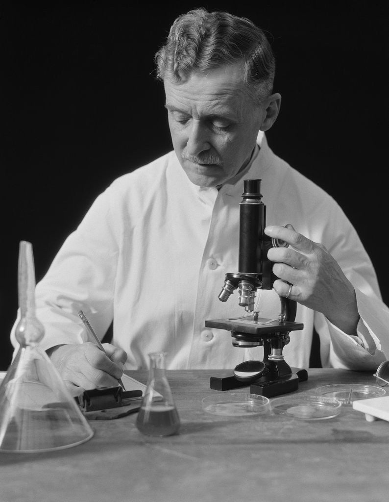 Detail of Male scientist wearing white lab coat writing data on clip board holding focus control of microscope by Anonymous