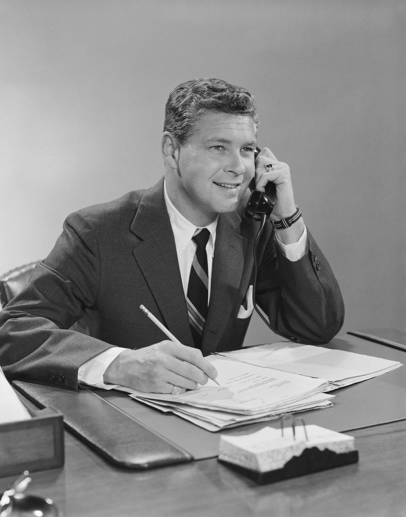 Detail of Businessman seated at desk talking on telephone smiling writing with pencil by Anonymous