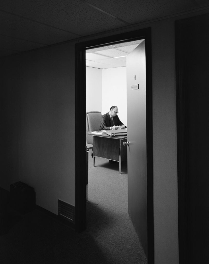 Detail of Executive businessman at desk seen working late through door ajar by Anonymous