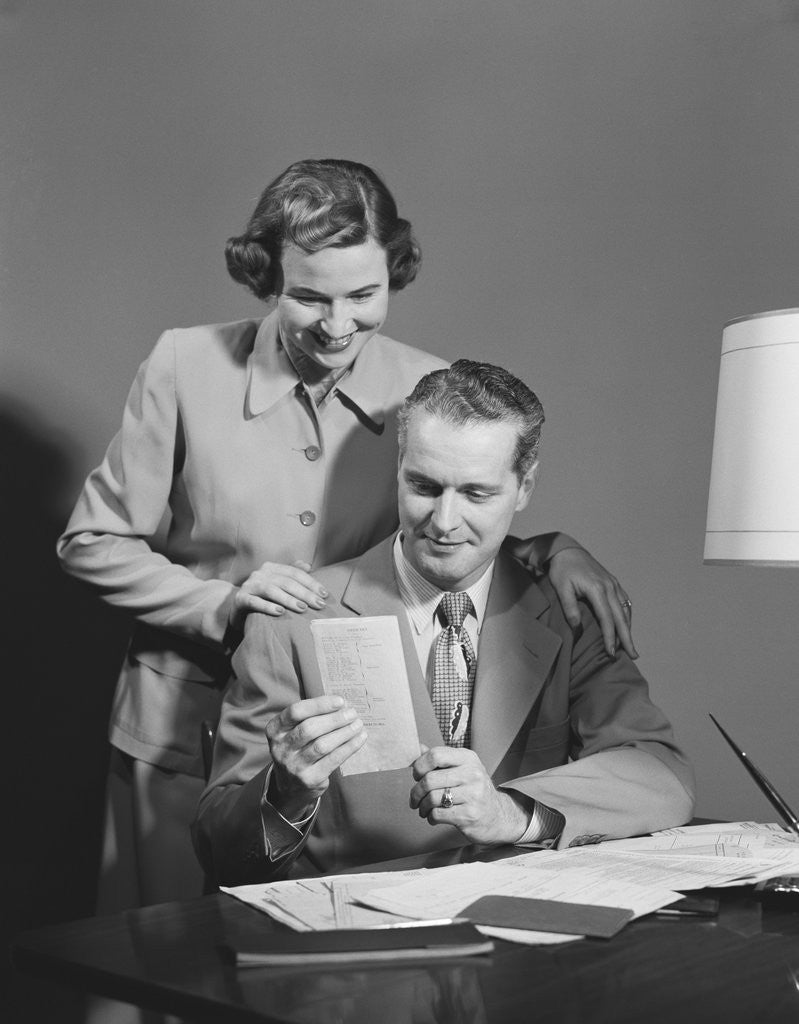 Detail of Couple man seated at desk holding paper woman standing looking over his shoulder smiling happy by Anonymous