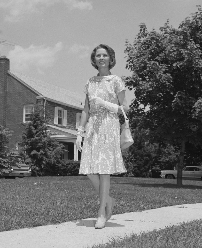 Woman wearing print dress holding handbag walking down suburban street by Corbis