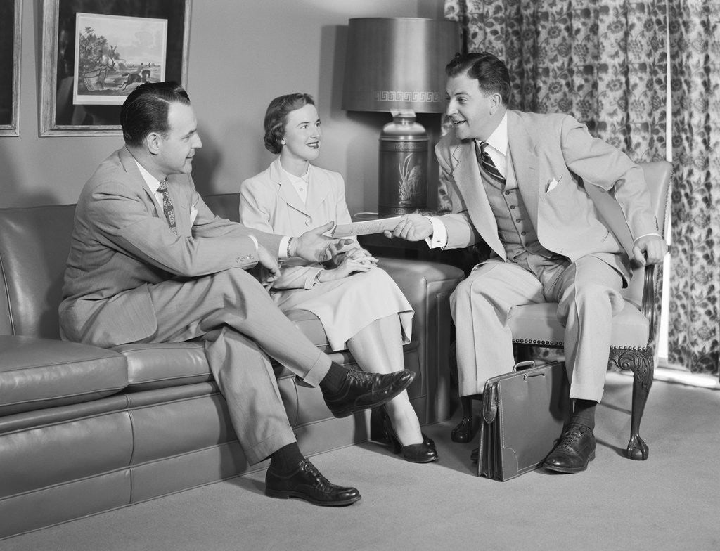 Detail of Couple sitting on couch talking to smiling insurance salesman handing them a policy by Corbis