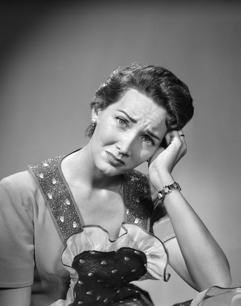 Detail of 1950s housewife woman in print apron hand up to temple headache pain sad depressed facial expression worried serious looking at camera by Anonymous