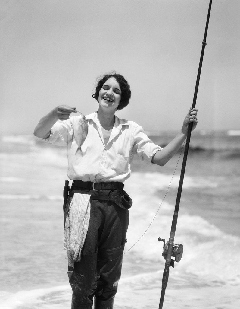 Detail of 1920s 1930s smiling woman standing in ocean surf wearing rubber waders holding fish and fly fishing rod by Anonymous