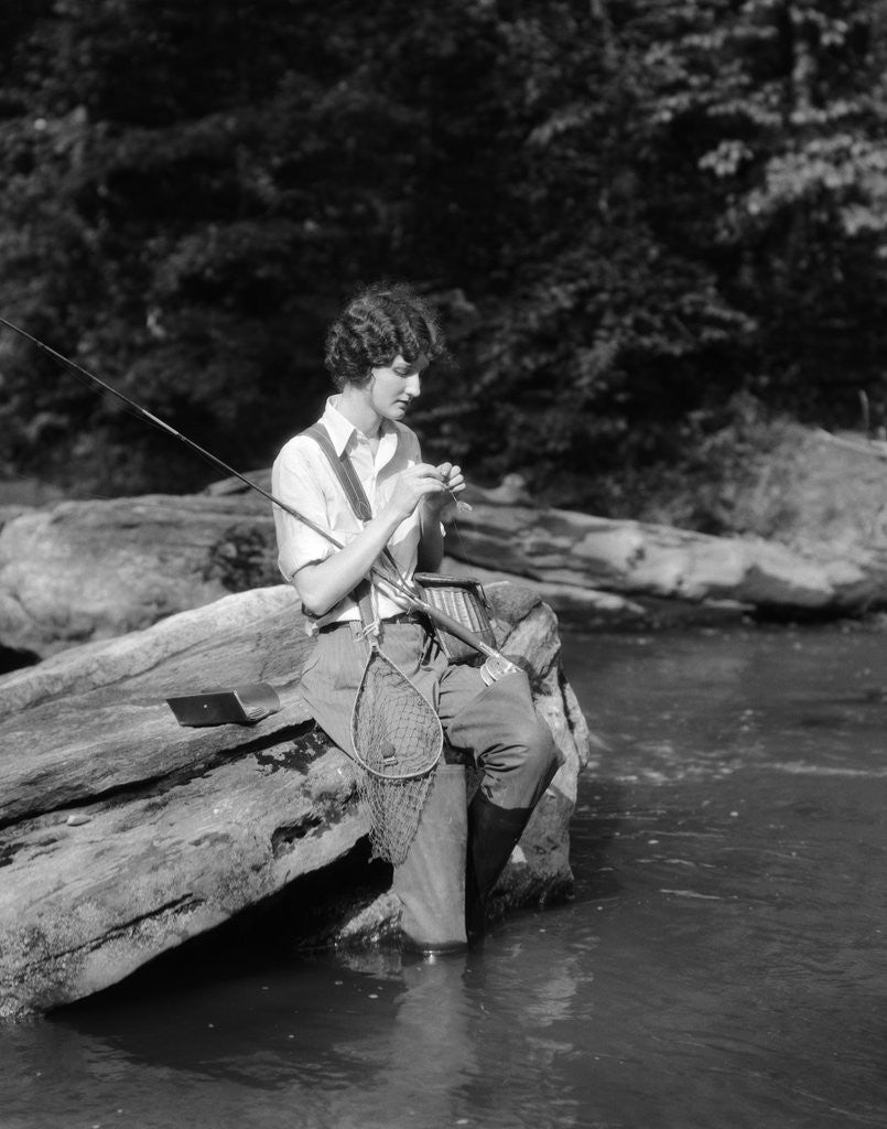 Detail of 1920s 1930s woman sitting on rock by side of stream wearing waders putting fly on tippet by Anonymous