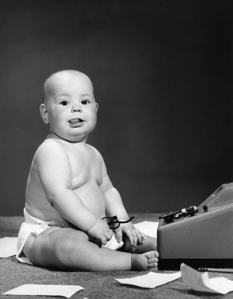Detail of 1950s 1960s baby in diaper sticking out tongue holding glasses sitting before adding machine looking at camera by Anonymous