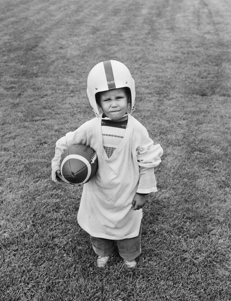 Detail of 1950s boy standing in grass wearing oversized shirt & helmet holding football by Anonymous