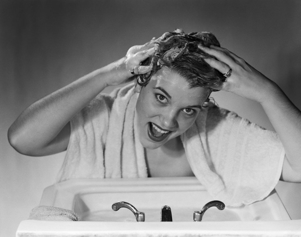 Detail of 1950s smiling woman washing shampooing hair in sink looking at camera by Anonymous