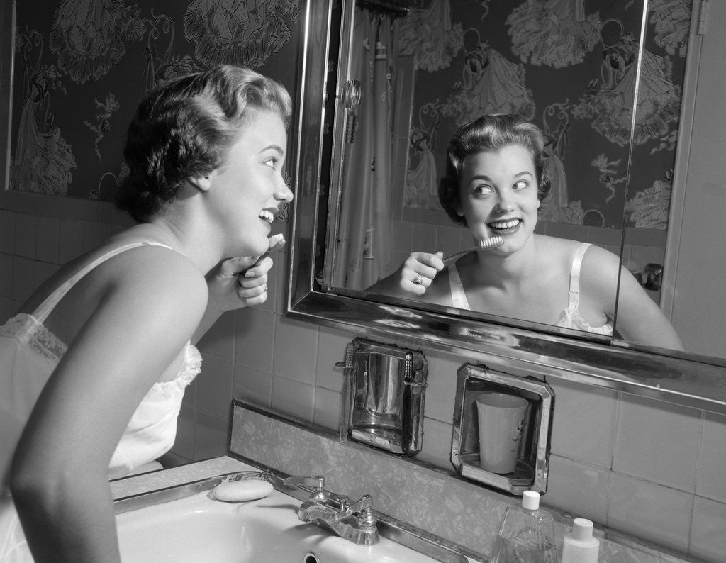 Detail of 1950s smiling woman looking into bathroom mirror brushing teeth by Anonymous