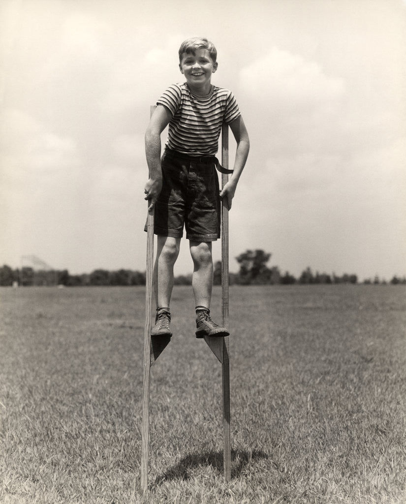 Detail of 1930s 1940s smiling happy boy wearing striped shirt & short pants walking on pair of stilts looking at camera by Anonymous