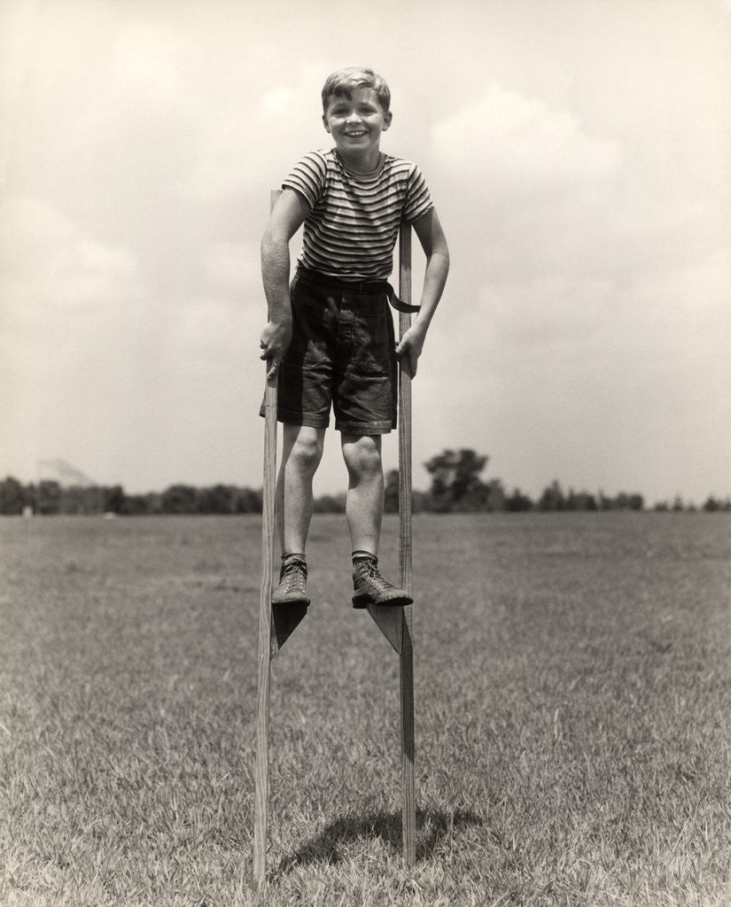 Detail of 1930s 1940s smiling happy boy wearing striped shirt & short pants walking on pair of stilts looking at camera by Corbis