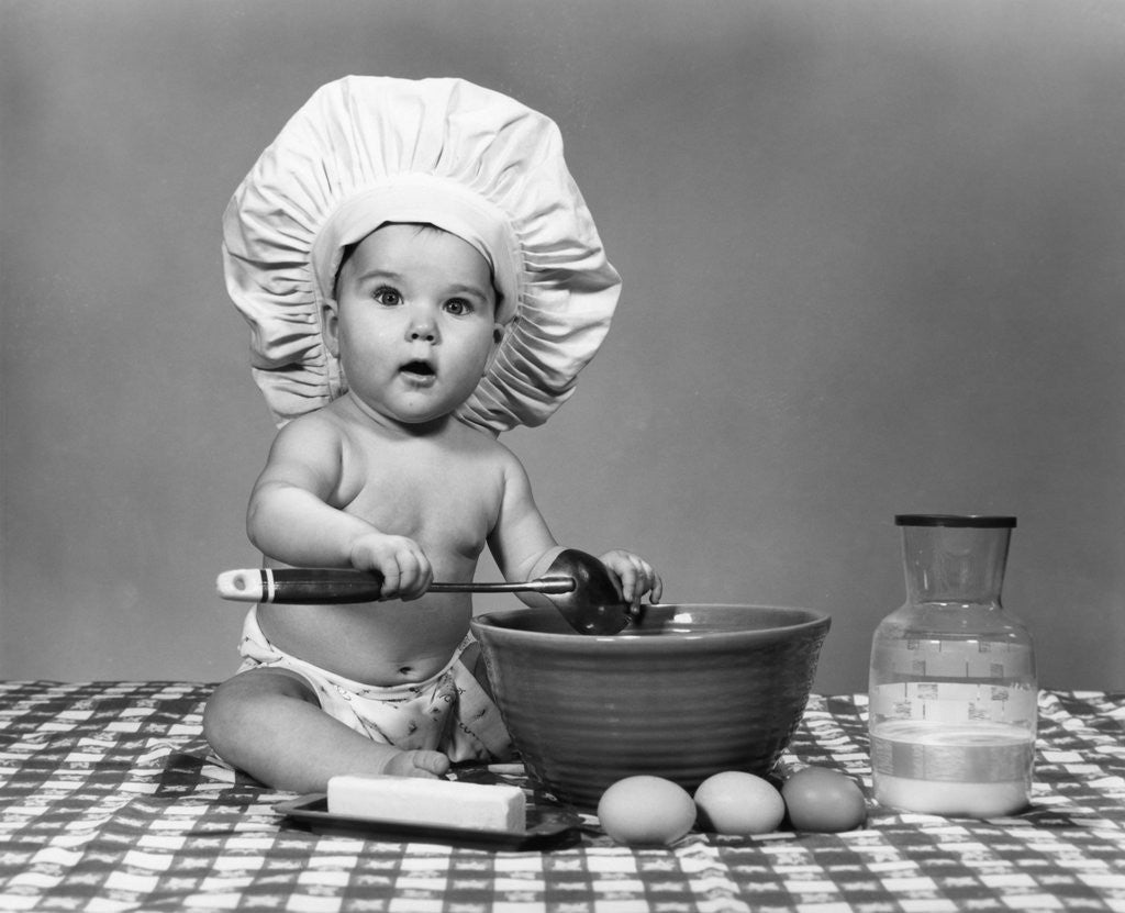 1960s baby seated on checkered tablecloth wearing chef's hat mixing eggs milk & flour in large bowl looking at camera by Corbis