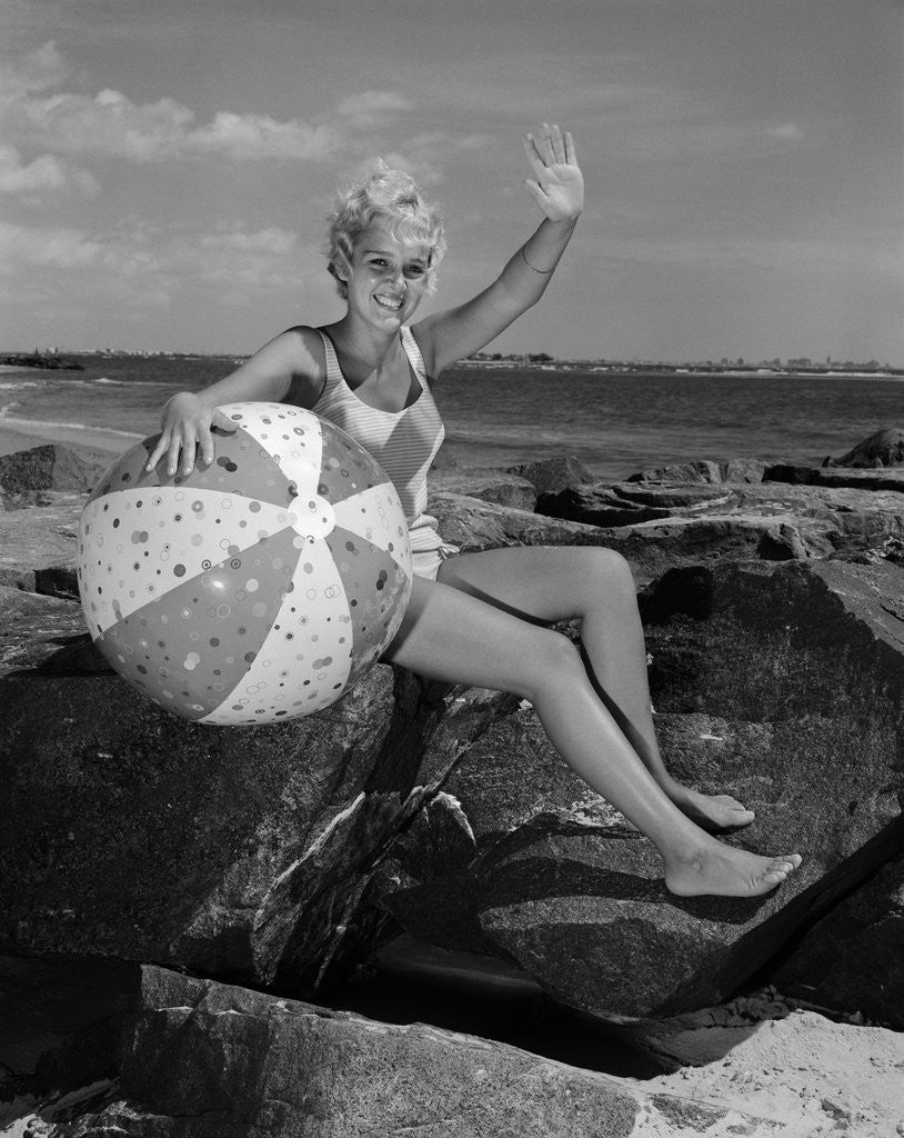 Detail of 1960s smiling blond teenage girl sitting on rocky shore holding a beach ball waving looking at camera by Anonymous